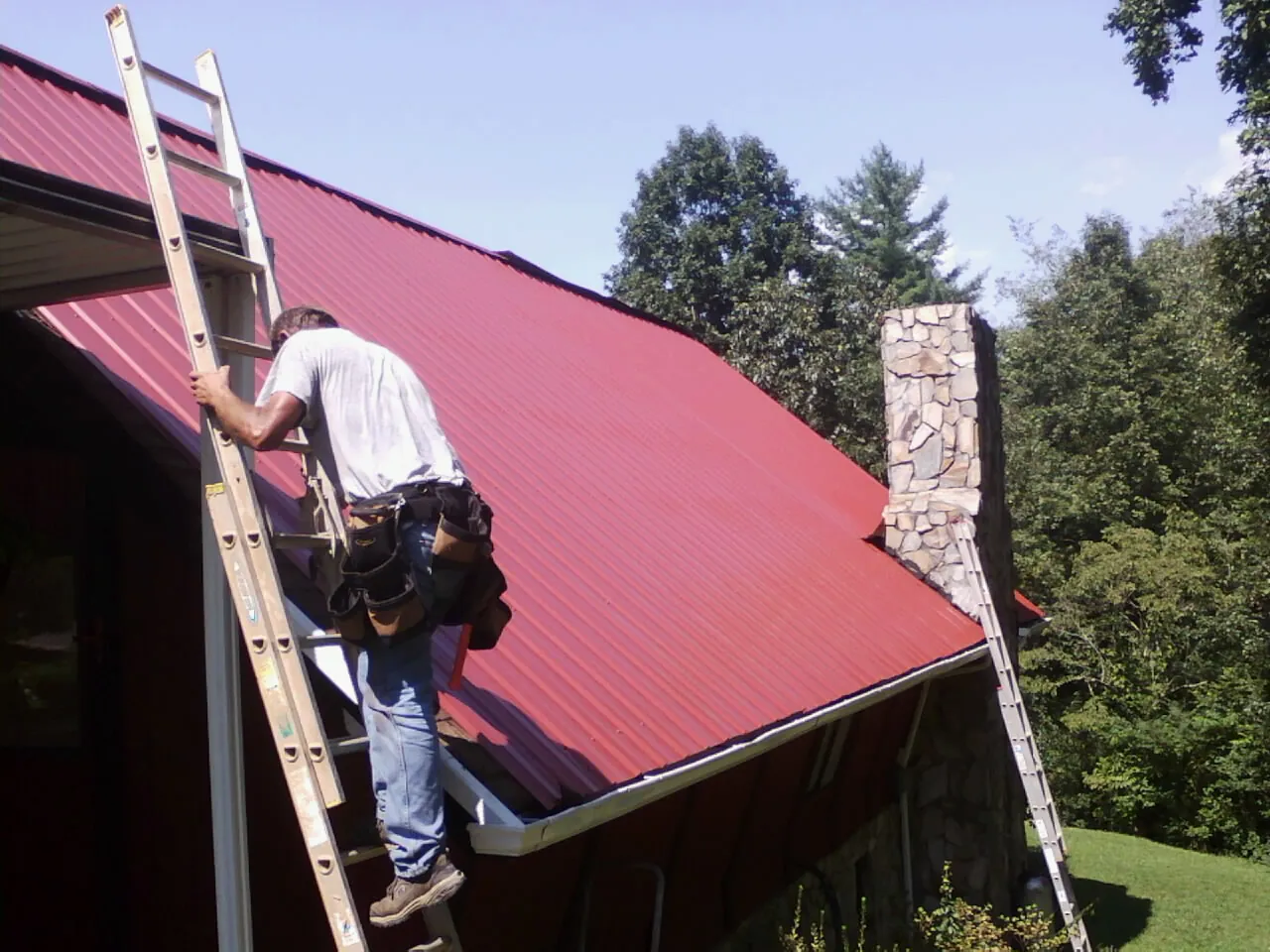 Roofer on a ladder beside a red metal roof and stone chimney — Soffit & Fascia Repair by Roofing Brothers in Hopkins